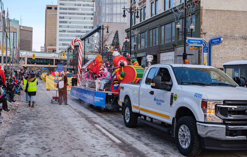 Un camion de Manitoba Hydro tire un char allégorique transportant une canne à sucre, des arbres de Noël et des personnages en costume. Un renne marche à côté du camion en recueillant des objets.