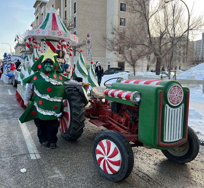 Une adulte déguisée en arbre de Noël décoré se tient devant un char allégorique à côté d’un tracteur.