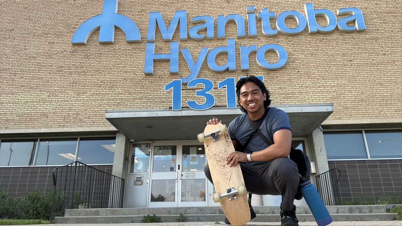 Homme accroupi qui tient une planche à roulettes devant un immeuble de bureaux de Manitoba Hydro.