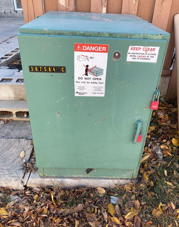 A squat green metal box on a concrete platform surrounded by grass and leaves. There’s a serial number on the top left of the box, a bright red DANGER sticker in the top middle, and a KEEP CLEAR sticker on the top right. The left of the box has two red locks.