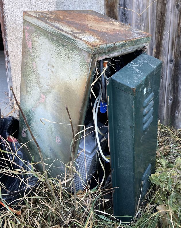 A photo of a knee-height green metal box among long grass and beside a fence. The box is rusty and leaning to one side; the right panel has vents on the top and bottom and is separated from the rest of it. There are wires coming out of the box with diameters no bigger than a pencil.