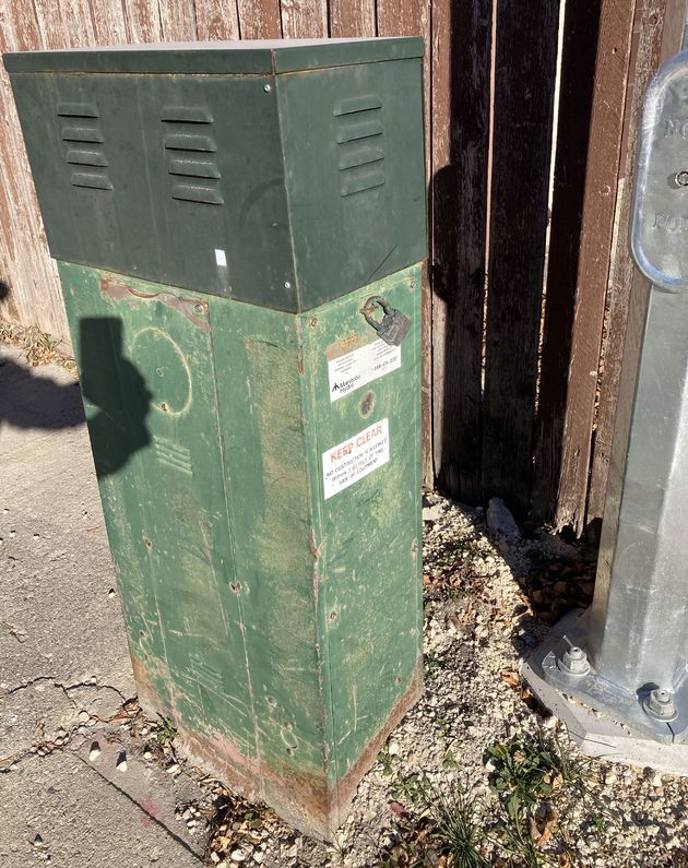  A photo of a waist-high green metal box with two shades of green. The top third, a forest green colour, has three vents visible. The bottom two thirds of the box, a faded and rusty grass colour, have no vents. Near the middle of the box is a metal lock, a “Manitoba Hydro” sticker, and a “KEEP CLEAR” sticker below it.